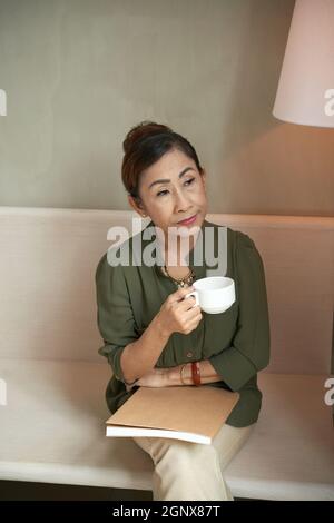 Pensive senior Asian lady in green blouse sitting on sofa in cafe and holding book on knees while drinking coffee Stock Photo