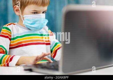 Boy in medical face protection mask indoors on blue background. Child sitting at the table and studying on the laptop. Stock Photo