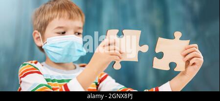 Boy in medical face protection mask indoors on blue background with two pieces of puzzle. Sick child. Coronavirus safety and allergy concept Stock Photo