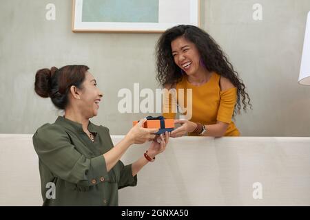 Cheerful young woman with curly hair giving present to senior mother while congratulating her on birthday Stock Photo