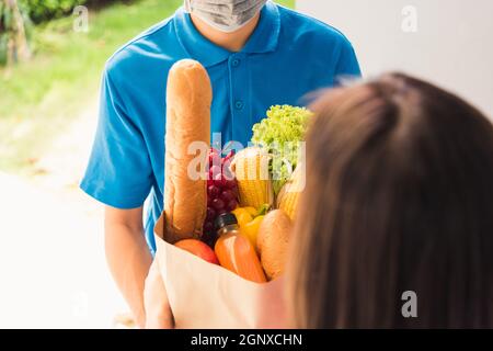 Asian young delivery man in uniform wear protective face mask he making grocery service giving fresh food to woman customer receiving front house unde Stock Photo