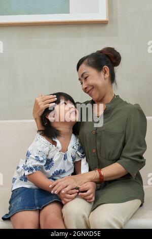 Smiling loving Asian grandma with hair bun sitting on sofa and stroking granddaughter while cuddling her Stock Photo