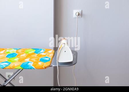 Electric iron plugged into an outlet on an ironing board against a gray wall. Ironing clothes. Stock Photo
