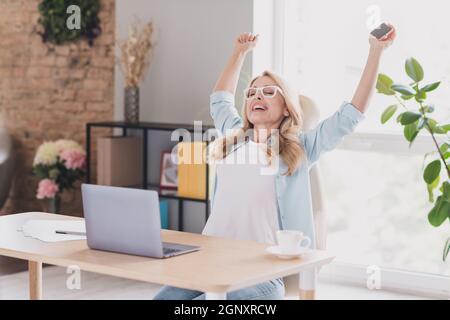 Photo of astonished funny age woman dressed blue shirt spectacles sitting table rising fists chatting modern gadget indoors home room Stock Photo