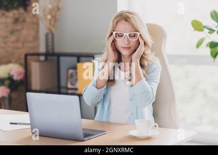 Photo of tired upset mature lady wear blue shirt glasses sitting table typing modern device arms temples indoors house room Stock Photo