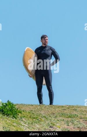 A male surfer carrying his surfboard and looking out to sea at Fistral in Newquay in Cornwall. Stock Photo