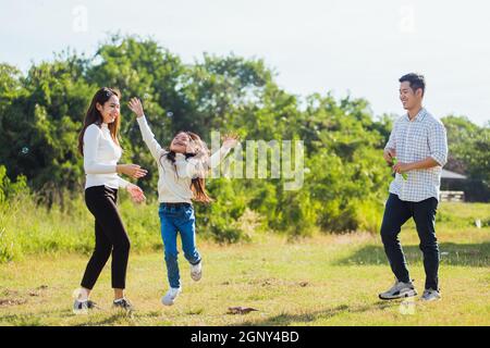 Happy Asian lifestyle family mother, father and little cute girl child having fun together and enjoying outdoor play blowing soap bubbles in the garde Stock Photo