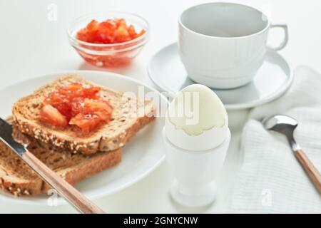 Breakfast. Still life from boiled egg, appetizing  toasts with apple jam and cup of coffee on white background. Selective focus on boiled egg Stock Photo