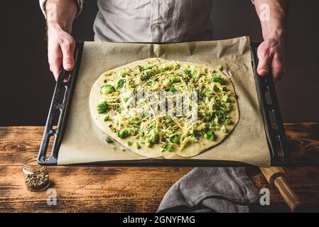 Man holding baking dish with uncooked pizza with broccoli, pesto sauce, spices and cheese Stock Photo