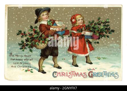 Original embossed Edwardian era Christmas greetings postcard of cute young children wearing winter clothes, carrying gifts and holly, snowy scene, published by International Art Publishing Co. Ltd. printed in Germany circa 1910 Stock Photo