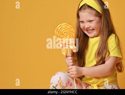 Happy smiling, sly kid girl in yellow t-shirt and colorful leggings sits squatting looking at big lollipop she holds Stock Photo