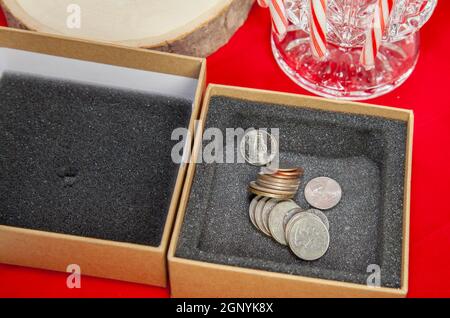 Empty, unwrapped gift box filled with pennies, nickels, dimes, quarters, and a dollar coin, on a red table top with candy canes in the background Stock Photo
