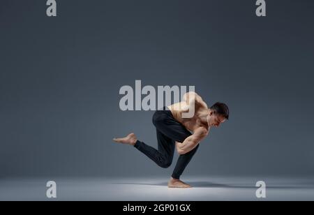 Male yoga keeps balanc in a difficult pose, grey background. Strong man doing yogi exercise, asana training, top concentration, healthy lifestyle Stock Photo