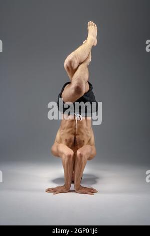 Male yoga standing on his head and hands, meditation, grey background. Strong man doing yogi exercise, top concentration Stock Photo