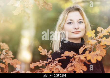 Blond girl is posing behind the branches of an oak in the autumn park. Horizontally. Stock Photo