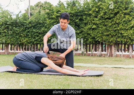 Asian man and woman training yoga outdoors in meditate pose sitting on green grass. Young couple practicing doing stretching in nature a field garden Stock Photo