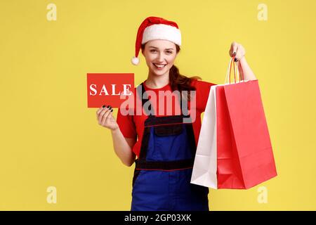 Optimistic delivery woman shows card with sale inscription and shopping bags, discounts for shipping, wearing blue overalls and santa claus hat. Indoor studio shot isolated on yellow background. Stock Photo