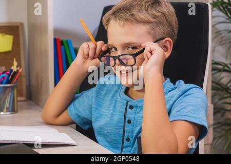 Home learning, online education. Schoolboy with glasses studies at home. Boy while sitting at table in front of laptop and looks at camera. Stock Photo