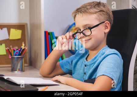 Home learning, online education. Schoolboy with glasses studies at home. Boy while sitting at table in front of laptop and looks at camera. Stock Photo