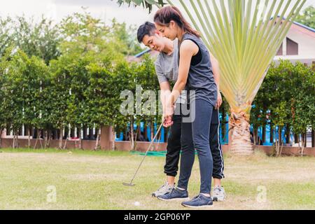 Asian young man support teaching training woman to play perfect golf while standing together in nature a field garden park. Couple trainer giving a le Stock Photo