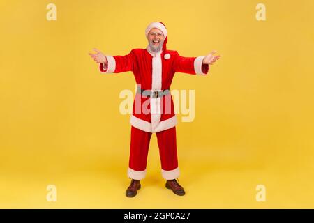 Full length of happy elderly man with gray beard wearing santa claus costume raising hands to embrace, welcoming and smiling delighted with meeting. Indoor studio shot isolated on yellow background. Stock Photo
