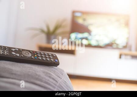 TV remote control in the foreground, tv in the blurry background. Streaming. Stock Photo