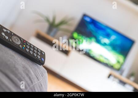 TV remote control in the foreground, tv in the blurry background. Streaming. Stock Photo