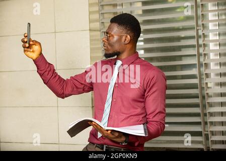 young busy African man making a gesture while talking while holding his notebook. A concerned man holding a document in his hand. Wearing a burgundy r Stock Photo
