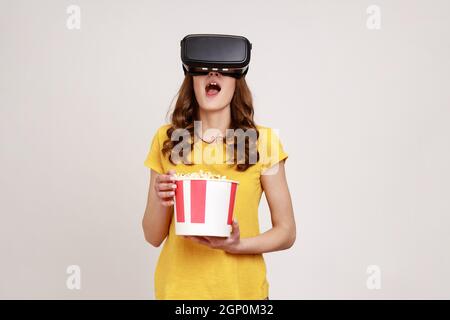 Young girl in yellow T-shirt getting experience VR headset, using augmented reality eyeglasses being in virtual reality, watching movie with popcorn. Indoor studio shot isolated on gray background. Stock Photo