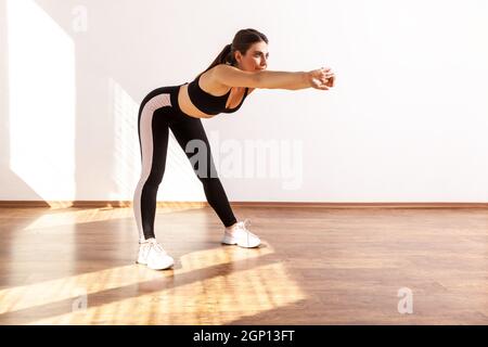 Woman doing sport, bending and stretching hands out, warming up training muscles for flexibility, wearing black sports top and tights. Full length studio shot illuminated by sunlight from window. Stock Photo