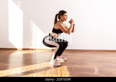 Full length portrait of woman doing split or lunge squat exercise at home or fitness gym, wearing black sports top and tights. Indoor studio shot illuminated by sunlight from window. Stock Photo