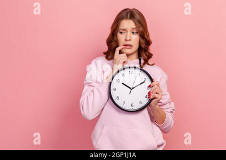 Portrait of impatient nervous teenage girl in hoodie biting her nails and holding big wall clock, deadline. Indoor studio shot isolated on pink background Stock Photo