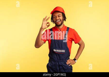 Portrait of sappy handyman with beard wearing overalls showing ok sign, being satisfied of done work in time, recommend service industry. Indoor studio shot isolated on yellow background. Stock Photo