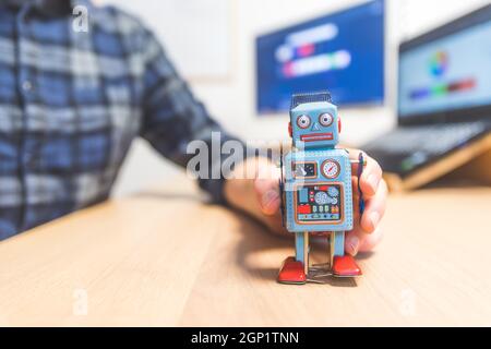 Toy robot on a desktop, man working on the computer in the background. Symbol for artificial intelligence, chatbots or social bots and algorithms Stock Photo
