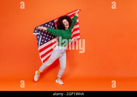 Full length photo of woman with Afro hairstyle wearing green casual style sweater raised arms, holding american flag, celebrating national holiday. Indoor studio shot isolated on orange background. Stock Photo
