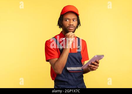 Portrait of pensive handyman wearing blue overalls standing with paper notebook in hands, writing down orders, having thoughtful expression. Indoor studio shot isolated on yellow background. Stock Photo