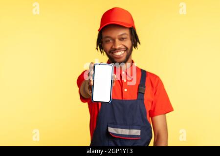 Good looking craftsman wearing blue uniform holding out showing mobile phone with empty display for advertisement online app for maintenance services. Indoor studio shot isolated on yellow background. Stock Photo