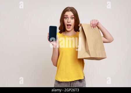 Shocked brown haired teenage girl in yellow T-shirt with opened mouth holding paper bags and smartphone with empty screen, online shopping app. Indoor studio shot isolated on gray background. Stock Photo