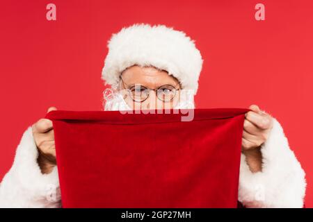 Santa claus in eyeglasses covering face with sack isolated on red Stock Photo
