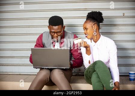 two young business couple sitting outdoors paying bills with credit card using a laptop while smiling Stock Photo