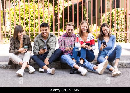 Multicultural friends laughing using smartphone sitting on the sidewalk in the city - Young people addicted by mobile smart phones - Technology concep Stock Photo