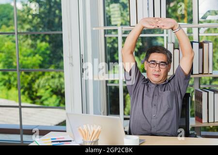 Asian businessman tired overworked he stretch oneself on the desk. senior man with eyeglasses break stretching his arms on table at his working place Stock Photo