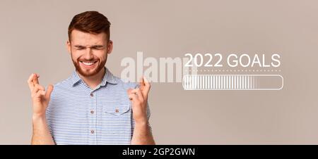 Motivated young red haired guy crossing fingers, setting goals for upcoming 2022 with closed eyes, making New Year resolution on grey studio backgroun Stock Photo