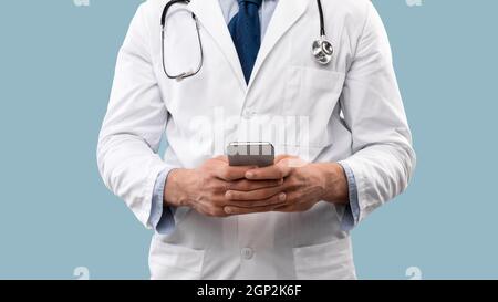 Cropped Shot Of Doctor Man Using Smartphone With Medical Application Standing In Studio Over Blue Background. Unrecognizable Professional Physician Po Stock Photo