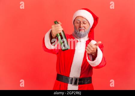 Funny elderly man with gray beard wearing santa claus costume standing with beer bottle, being drunk, standing with closed eyes and showing thumb up. Indoor studio shot isolated on red background. Stock Photo