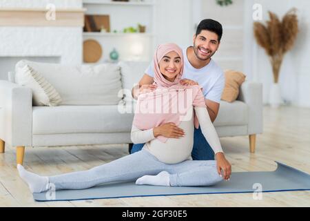 Pregnancy workout. Happy arab spouses exercising together at home, young expectant woman stretching legs while man massaging wife's shoulders and smil Stock Photo