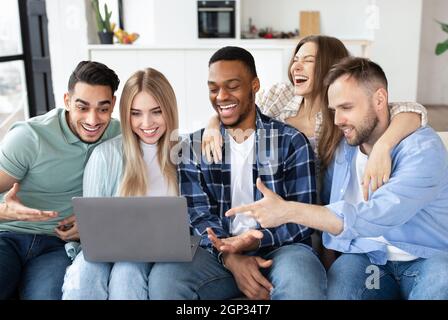 Group of multiracial millennial friends using laptop at home, chatting online, having fun together. Diverse young people communicating on web, video c Stock Photo