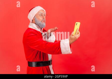 Side view of elderly man with gray beard wearing santa claus costume sending air kiss to followers while broadcasting livestream or making selfie. Indoor studio shot isolated on red background. Stock Photo