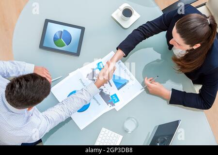 Top view of young business partners shaking hands on deal at desk in office.Documents and digital tablet on table showing statistics and graphics. Stock Photo