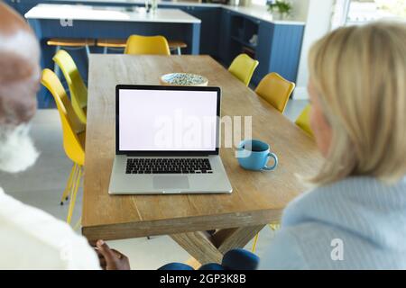 Happy senior diverse couple in kitchen sitting at table, using laptop Stock Photo